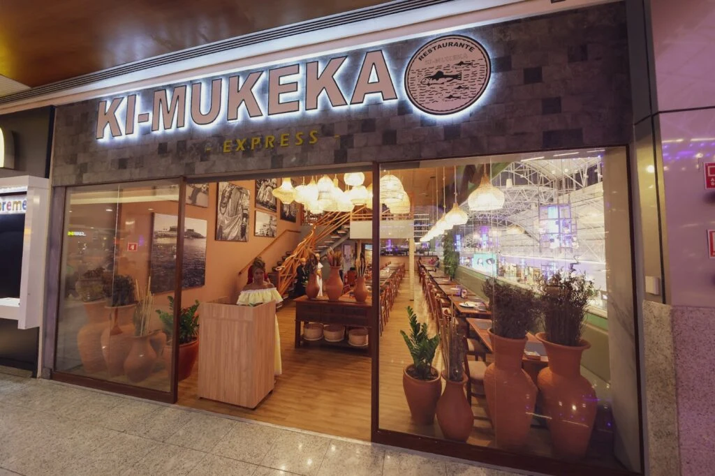 Fachada de restaurante em um shopping, com o nome "KI-MUKEKA" iluminado acima da entrada e mesas visíveis no interior.